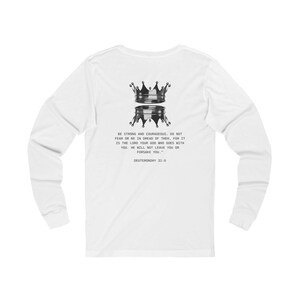 May include: White long-sleeve shirt featuring a two-tiered crown design in black. The shirt includes the text "BE STRONG AND COURAGEOUS, DO NOT FEAR OR BE IN DREAD OF THEM, FOR IT IS THE LORD YOUR GOD WHO GOES WITH YOU. HE WILL NOT LEAVE YOU OR FORSAKE YOU. DEUTERONOMY 31:6."