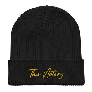 Op de afbeelding: Zwarte gebreide beanie met een geborduurd logo in goud met de tekst "The Notary".