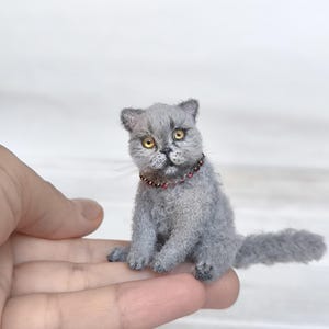 May include: A miniature needle-felted grey cat figurine with textured fur, amber-coloured eyes, and a small collar featuring tiny red beads. The cat is depicted in a seated position with its tail extended to the side, showcasing detailed craftsmanship.