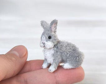 May include: A miniature gray and white rabbit figurine with detailed fur texture. The small bunny is perched on a finger, showcasing its delicate features and lifelike appearance. The rabbit has upright ears and a fluffy tail.