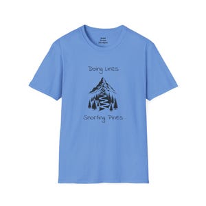 May include: A light blue t-shirt with a black mountain and pine tree graphic. The text "Doing Lines" is above the mountain, and "Snorting Pines" is below. The brand name "Bald Crow Designs" is printed at the top.