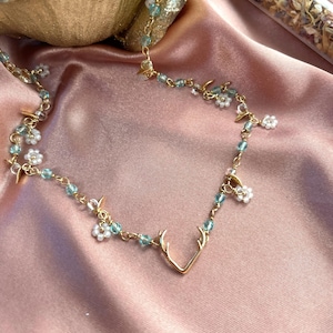 May include: A gold-toned necklace featuring a central deer antler charm, interspersed with small white beaded flower motifs and translucent light blue crystal beads on a delicate gold-coloured metal chain link structure.
