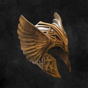 May include: A gold-colored helmet with intricate wing and knot designs. The helmet features a pointed front and a detailed, textured surface. The wings extend upwards and outwards, adding to the helmet's ornate appearance. The helmet is likely for cosplay or display.