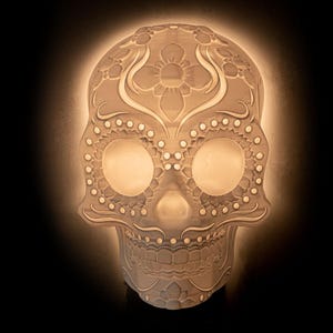 May include: White ceramic sugar skull lamp with intricate floral and dot designs. The lamp is lit and glowing.