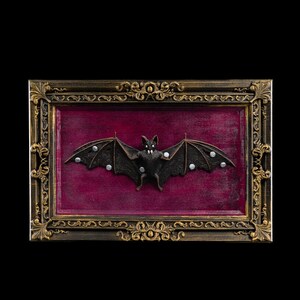 May include: A framed, dark-colored bat sculpture with outstretched wings and silver accents. The bat is set against a deep red background within an ornate, gold-toned frame. The frame has intricate detailing.