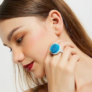 May include: A blue and white ring with a textured, brush-like surface. The ring is worn on a person's finger.