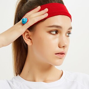 May include: A woman with long brown hair wearing a red headband and a white t-shirt. She is wearing a blue and red ring on her finger.