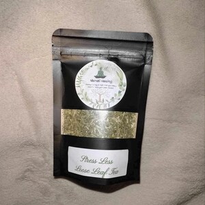 May include: A black resealable pouch with a white label that reads "Stress Less Loose Leaf Tea". The pouch contains dried green tea leaves. The label also features a logo with the text "Maraki Healing".