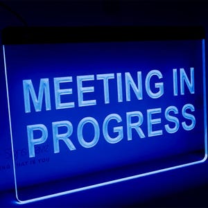 Può includere: Un cartello rettangolare illuminato di blu con la scritta bianca "MEETING IN PROGRESS". Il cartello ha un bordo nero nella parte superiore e un logo nell'angolo in basso a sinistra.