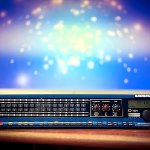 May include: A blue Samson D-3500 audio processor with various buttons, knobs, and a digital display. The rack-mounted device is positioned in front of a computer monitor with a blurred blue and white background.