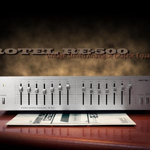 May include: A silver ROTEL RE-500 vintage stereo graphic equalizer with multiple sliders and control buttons. The unit has the text "ROTEL RE-500 Vintage Stereo Analogue Graphic Equalizer" printed on the front. A manual rests below the unit.