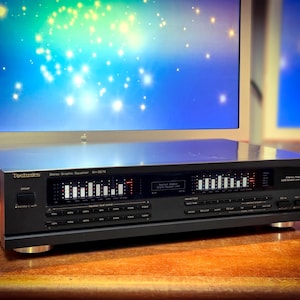 May include: A black Technics SH-GE70 stereo graphic equaliser with illuminated display bars. The unit has multiple buttons and a power switch. The background features a computer monitor with a blue and green gradient.