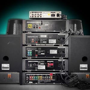 Puede incluir: Una pila de equipos de audio negros, que incluyen altavoces, un reproductor de CD y un receptor, con varios puertos de entrada y salida visibles. Los cables conectan los componentes. La configuraci&oacute;n est&aacute; contra un fondo verde oscuro.