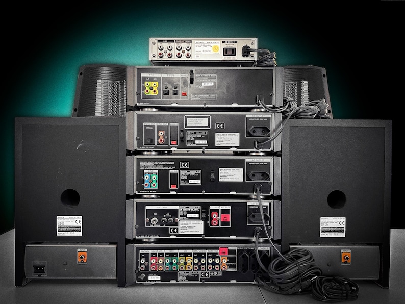 Puede incluir: Una pila de equipos de audio negros, que incluyen altavoces, un reproductor de CD y un receptor, con varios puertos de entrada y salida visibles. Los cables conectan los componentes. La configuraci&oacute;n est&aacute; contra un fondo verde oscuro.