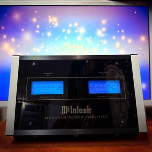 Puede incluir: Amplificador de potencia McIntosh MCC404M negro con dos pantallas azules iluminadas. El amplificador tiene detalles plateados y está colocado frente a un monitor de ordenador que muestra un diseño estrellado azul y blanco.