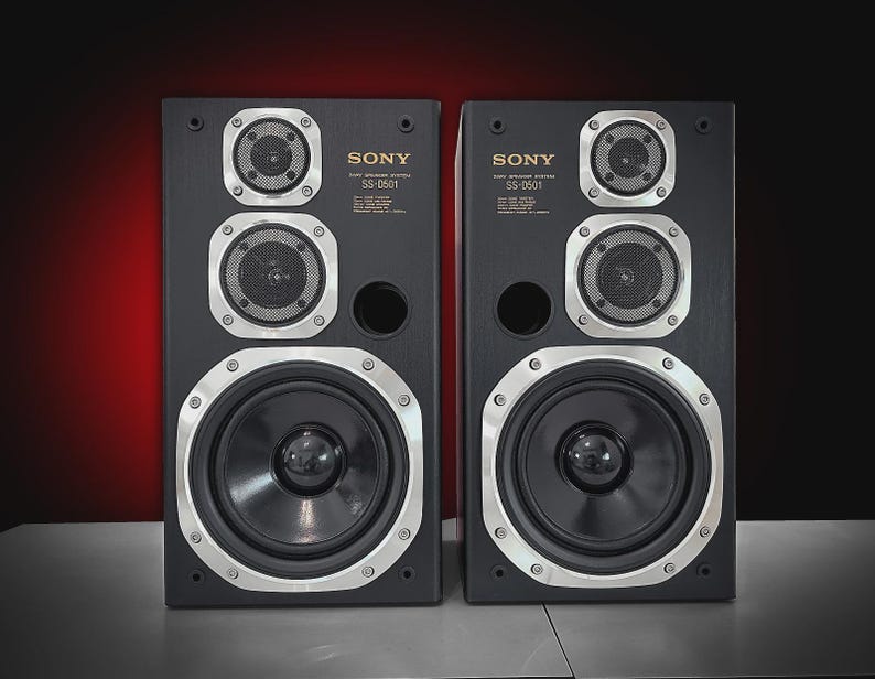 Puede incluir: Dos altavoces negros Sony SS-0501 con detalles plateados. Cada altavoz tiene un woofer central grande, dos altavoces m&aacute;s peque&ntilde;os y un puerto circular. Fondo degradado de rojo oscuro a negro.