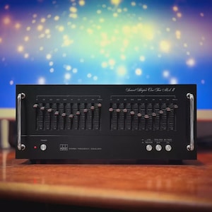 May include: A black audio equalizer with multiple sliders and knobs. The device has silver handles and the text "Sound Shaper One Ten Mk II" is printed on the top. The device is on a wooden surface with a blurred background.