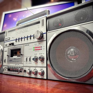 May include: Silver vintage boombox with large speakers and a cassette player. The boombox has multiple dials, buttons, and a handle. The speakers are covered with a mesh grill. The boombox is sitting on a wooden surface.
