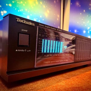 May include: A black Technics stereo graphic equaliser SH-8046. The device has a power button, a standby/on button, and a display showing blue vertical bars. The text "Technics" is visible on the top left.
