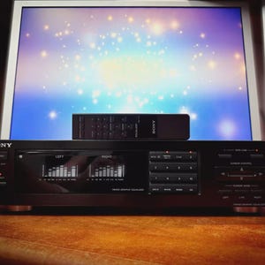 May include: A black Sony graphic equaliser with a remote control on top. The unit displays audio levels and has control buttons. The sides have a wood-grain finish. It is on a wooden surface. The unit measures approximately 43 cm wide.