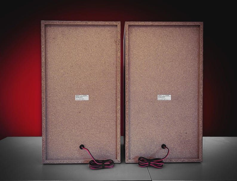 Puede incluir: Dos altavoces rectangulares con marco y parte trasera de madera marr&oacute;n claro. Cada altavoz tiene una peque&ntilde;a etiqueta blanca y un cable negro y rosa. Los altavoces est&aacute;n sobre un fondo degradado rojo y negro.