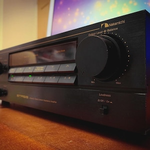 以下が含まれることがあります： 黒いNakamichiステレオアンプ。大きなコントロールノブと複数のボタンがあります。アンプにはディスプレイ画面があり、Nakamichiのロゴが見えます。ユニットは木製の表面にあります。