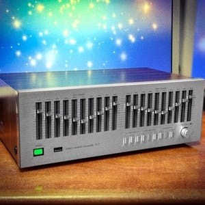 May include: A silver stereo graphic equaliser with multiple sliders and control knobs. The device has a brushed metal finish and a green power indicator light. The brand name and model number are visible on the front panel. The equaliser is on a wooden surface.