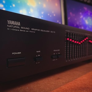 Può includere: Un equalizzatore grafico Yamaha Natural Sound EQ-70 nero. Il pannello frontale presenta il testo "YAMAHA", "NATURAL SOUND GRAPHIC EQUALIZER EQ-70" e pulsanti di controllo. Le luci rosse indicano i livelli audio.