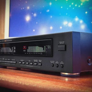 May include: A black audio equalizer with a digital display showing green and red bars. The device has several buttons and knobs, including "Equalizer" and "Memory". It sits on a wooden surface with a blue and white background.