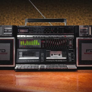 Puede incluir: Un radiocasete negro y plateado vintage con antena retráctil, reproductores de casete y controles de ecualizador. Los altavoces tienen detalles en rojo y negro con las letras "FD" visibles. El radiocasete está sobre una superficie de madera.