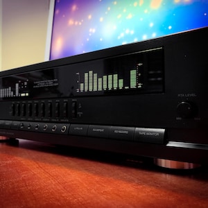 Puede incluir: Ecualizador de audio negro con una pantalla que muestra barras verdes. El dispositivo tiene varios botones y perillas, incluyendo "EQ Defeat" y "Tape Monitor". La unidad está sobre una superficie de madera.