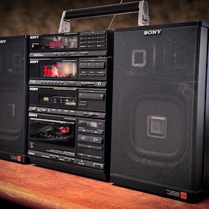 以下が含まれることがあります： シルバーのハンドルが付いた、黒いソニーのヴィンテージブームボックス。カセットプレーヤー、ラジオ、スピーカーを搭載。ブームボックスには、ラジオの周波数が表示されるデジタルディスプレイがあります。スピーカーは黒いメッシュで覆われています。