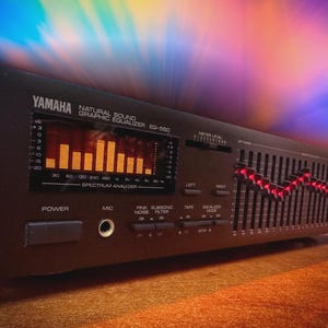 May include: A black Yamaha Natural Sound Graphic Equalizer EG-550. It has a spectrum analyzer display with orange bars and sliders with red lights. The front panel includes power and mic controls. A vintage audio component.