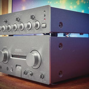 Puede incluir: Dos componentes de audio Kenwood plateados apilados. La unidad superior tiene múltiples perillas y botones, mientras que la unidad inferior presenta dos diales grandes. Las unidades están sobre una superficie de madera.