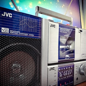 May include: A vintage JVC PC-100 detachable portable component system boombox. The silver and blue stereo features a speaker, cassette deck, and radio dial. The boombox has a handle for easy carrying.