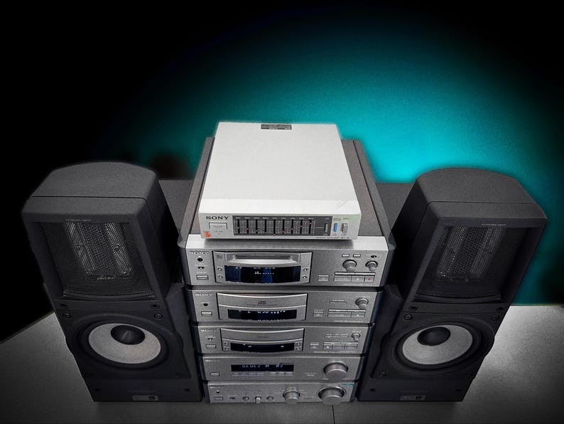 Puede incluir: Un sistema est&eacute;reo vintage con un ecualizador blanco y m&uacute;ltiples componentes plateados apilados. Dos grandes altavoces negros flanquean el sistema. La configuraci&oacute;n est&aacute; contra un fondo verde azulado oscuro.