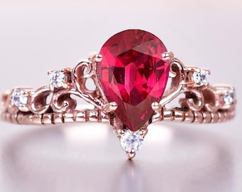 May include: A rose gold ring featuring a large, teardrop-shaped, deep red gemstone. The ring has a crown-like design with small, clear accent stones. The band is intricately detailed, giving it an elegant and ornate appearance.
