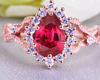 May include: A rose gold ring featuring a central, pear-cut, deep red gemstone, encircled by smaller white stones. The ring's band has delicate, swirling patterns. The ring is set against a pale grey backdrop, with white flowers subtly visible in the background.
