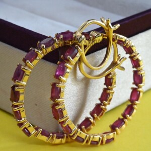May include: A pair of gold hoop earrings with rectangular, deep red gemstones set around the hoops. The earrings are displayed in an open jewelry box with a burgundy interior, set against a yellow background.