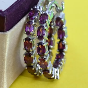 May include: Silver hoop earrings set with oval-cut, deep red gemstones. The earrings are shown on a yellow surface next to a beige and purple jewellery box. The gemstones are closely set.
