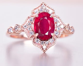 May include: Rose gold ring with a large oval ruby centre stone. The ruby is surrounded by smaller, clear gemstones and ornate filigree detailing. The ring's band is also adorned with small gemstones, creating a classic and elegant design.