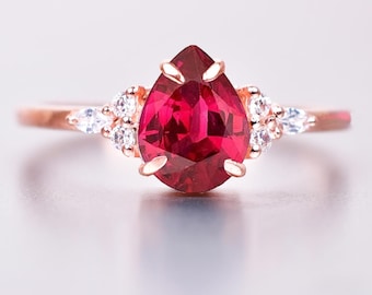 May include: Rose gold ring with a striking, pear-cut, deep red gemstone at its centre. The ring is embellished with smaller, clear gemstones flanking the main stone, offering a refined and elegant aesthetic. A beautiful piece of jewellery.