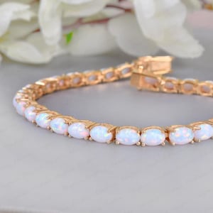 以下が含まれることがあります： 金色のブレスレット。楕円形のオパールのような宝石が連なっています。テニスブレスレットのデザインで、各宝石は互いに近くにセットされています。留め金が見え、明るい色の表面に表示されています。