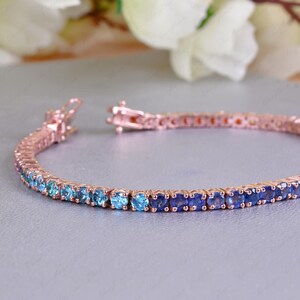 May include: Rose gold tennis bracelet with a gradient of blue and green gemstones. The bracelet has a secure clasp and is displayed on a light grey surface. The gemstones transition from light blue to dark blue and then to green.