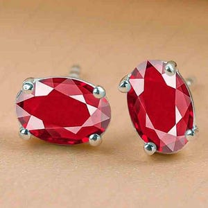 May include: A pair of stud earrings featuring oval-cut, faceted red gemstones. The gemstones are set in a silver-colored metal with four rounded prongs. The earrings are displayed on a neutral-toned surface.