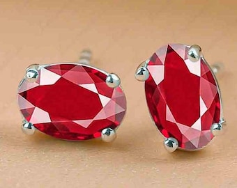 Peut inclure: Une paire de boucles d'oreilles à clous avec des pierres précieuses rouges taillées en ovale. Les pierres sont serties dans un métal argenté avec quatre griffes arrondies. Les boucles d'oreilles sont présentées sur une surface de couleur neutre.