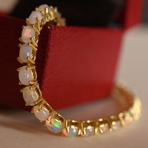 May include: A gold bracelet featuring oval-shaped opals. The opals are set in individual gold prongs, creating a continuous line of gemstones. The bracelet is displayed against a red background.
