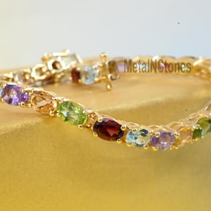 May include: A gold bracelet with a row of oval-shaped gemstones in various colors, including amethyst, aquamarine, peridot, citrine, and garnet. The bracelet is on a gold background.