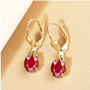 Könnte beinhalten: Ein Paar goldene Ohrringe mit roten Edelstein-Akzenten. Die Ohrringe haben ein schlichtes, elegantes Design und sind perfekt für den Alltag.