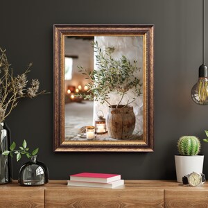 May include: A framed print of a still life featuring a rustic pot with a small olive branch, a lantern, and candles. The frame is brown with gold accents.